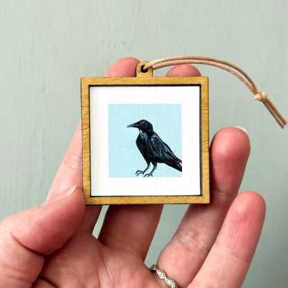 A person's hand is holding a small wooden frame containing a framed image of a black bird.