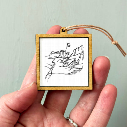 A hand holding a small wooden frame containing a black and white line drawing of a mountain landscape.