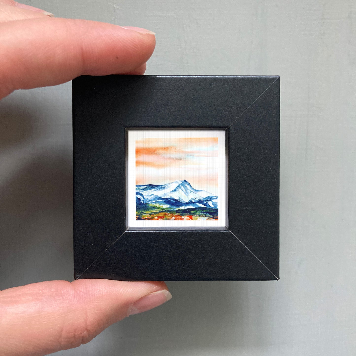 A person's hand is holding a small black frame containing a watercolor painting of a mountain landscape.