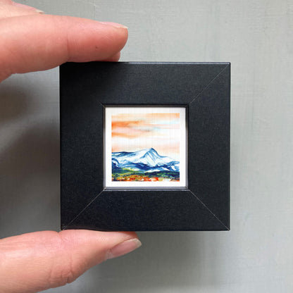 A person's hand is holding a small black frame containing a watercolor painting of a mountain landscape.