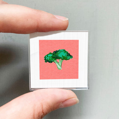 A person's hand is holding a small square with a picture of a green broccoli on it.