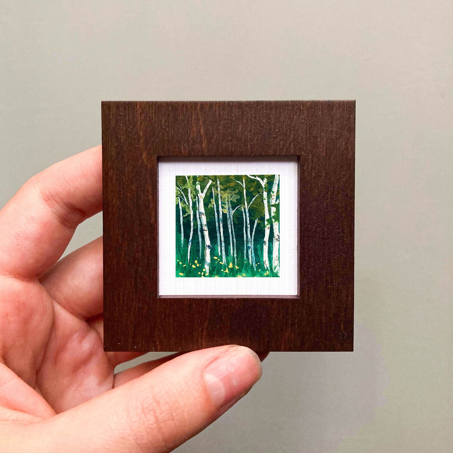 A hand is holding a small, square-shaped wooden frame containing a painting of a forest scene with white birch trees and green foliage.