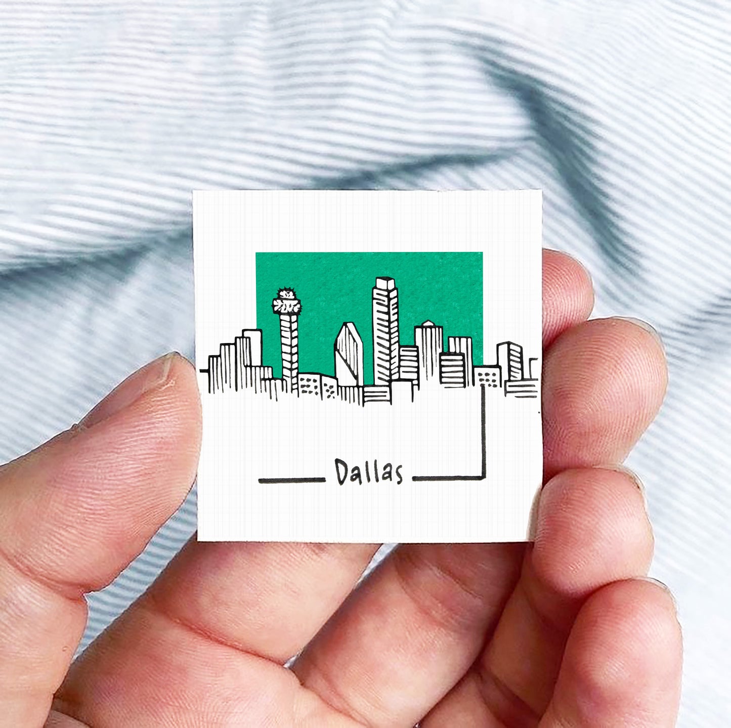 A hand holding a small square-shaped object with a drawing of a city skyline and the word "Dallas" written on it.