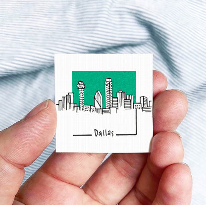 A hand holding a small square-shaped object with a drawing of a city skyline and the word "Dallas" written on it.