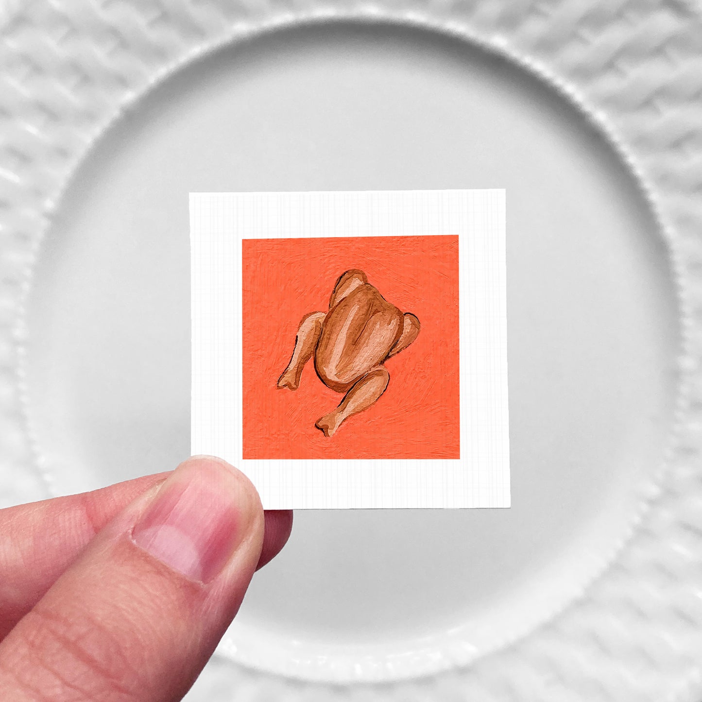 A hand is holding a small square with an illustration of a roasted chicken on it, against a white background.