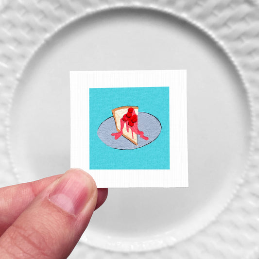 A hand is holding a small square with a picture of a slice of cheesecake on a blue background.