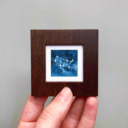 A hand is holding a small wooden frame containing a framed image of a starry night sky.