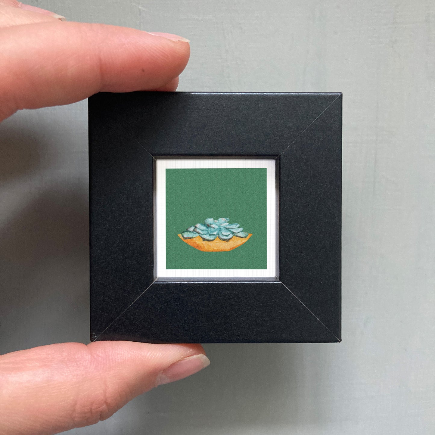 A hand is holding a small black frame containing a framed image of a green plant with blue flowers.
