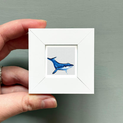 A hand is holding a small white picture frame containing a watercolor painting of a blue whale.
