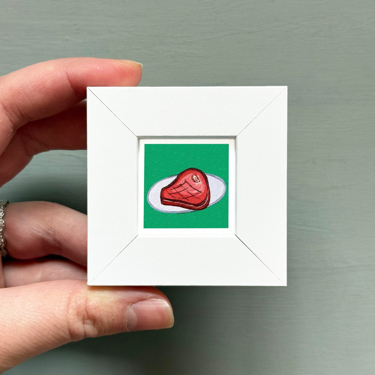 A hand is holding a small white square frame containing an illustration of a piece of meat on a green background.