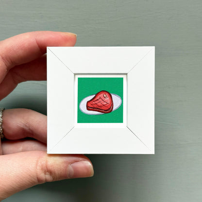 A hand is holding a small white square frame containing an illustration of a piece of meat on a green background.
