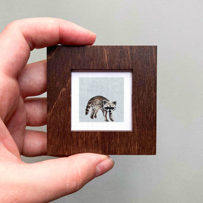 A hand is holding a small wooden frame containing a picture of a raccoon.