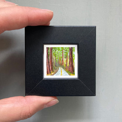 A person's hand holding a small black frame containing a miniature painting of a forest road with tall trees.