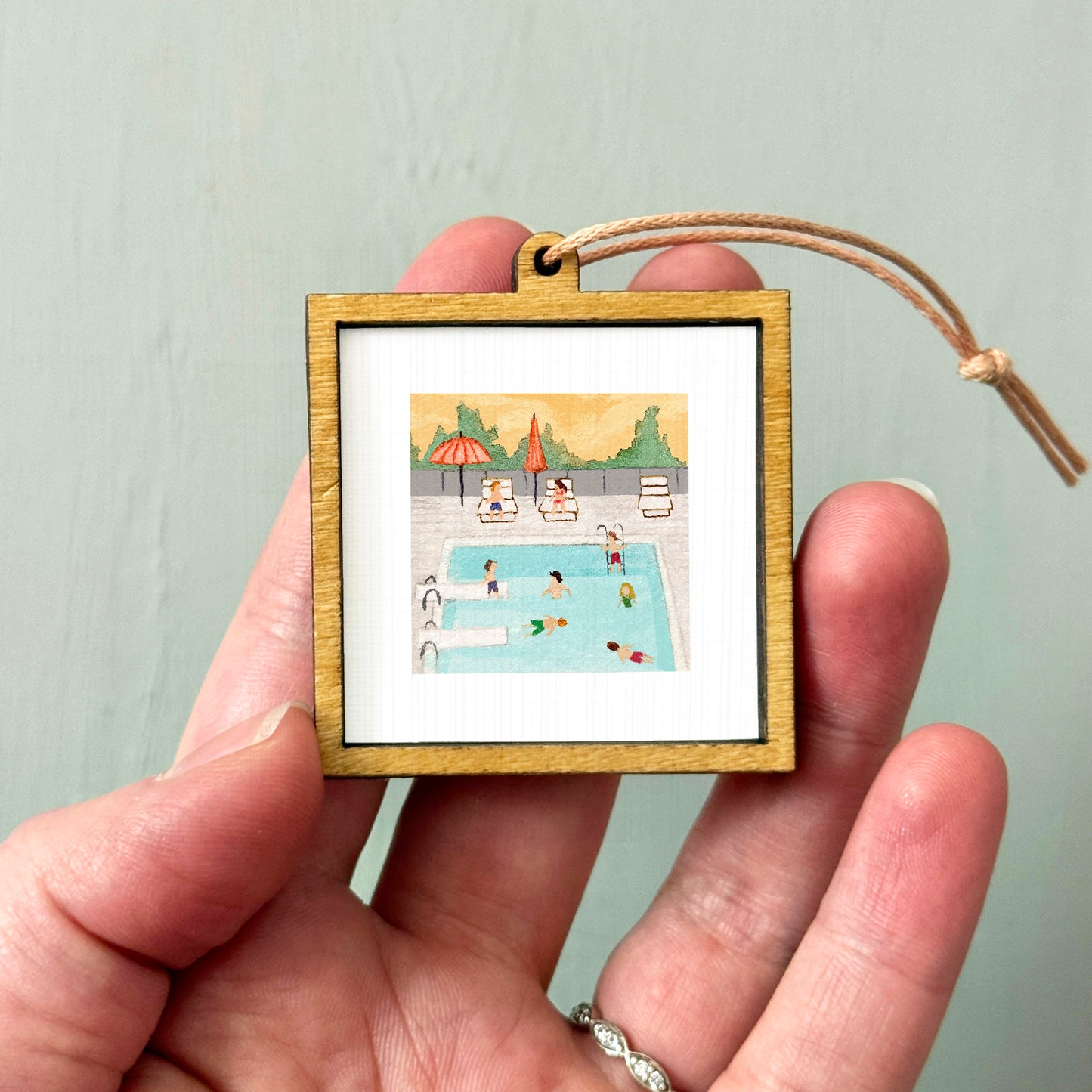 A hand holding a small wooden frame containing a miniature painting of a swimming pool with people enjoying the water.