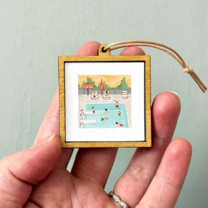 A hand holding a small wooden frame containing a miniature painting of a swimming pool with people enjoying the water.