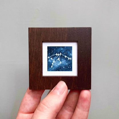 A hand is holding a small wooden frame containing a framed image of a starry night sky.
