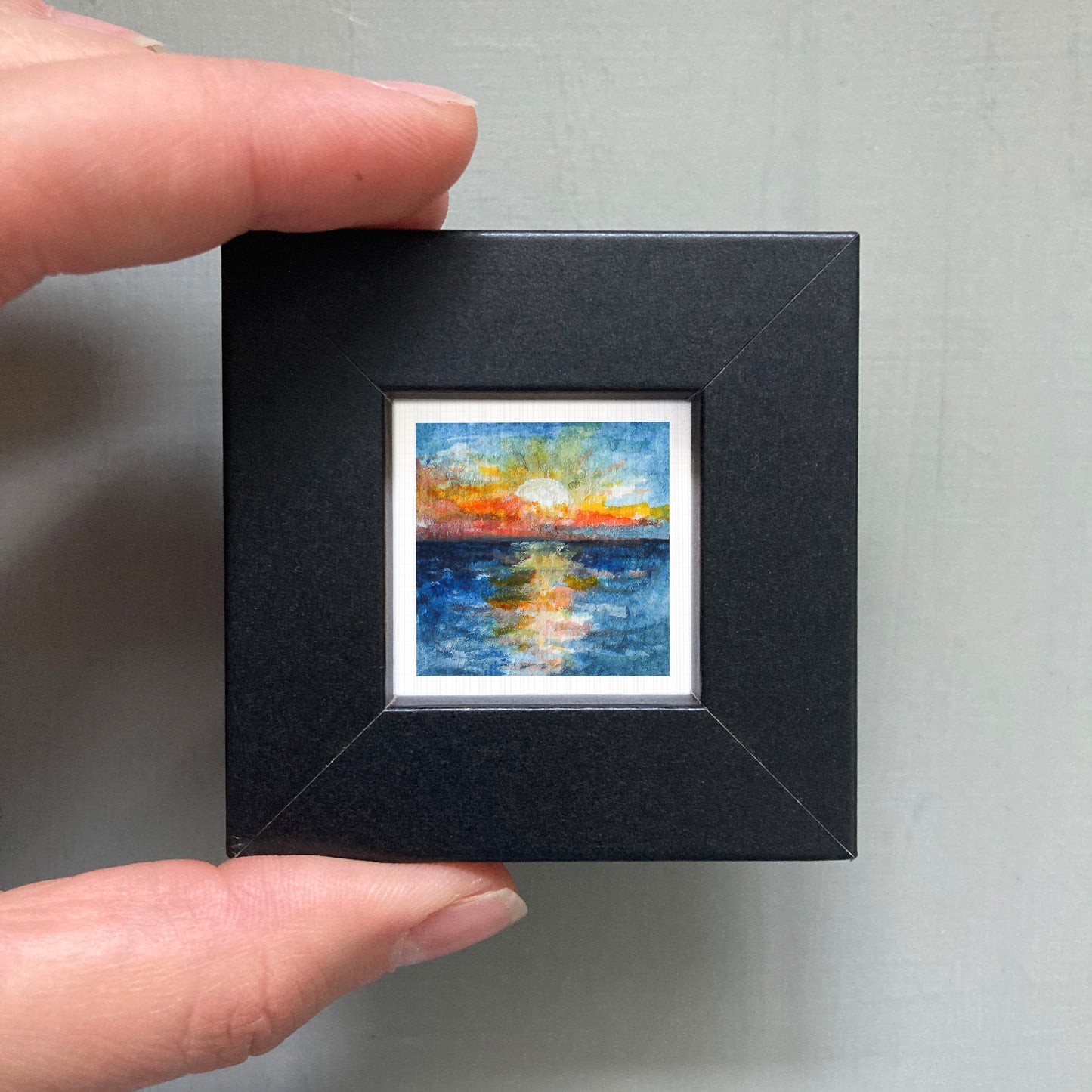 A person's hand holding a small, square-shaped black frame containing a miniature painting of a sunset over a body of water.