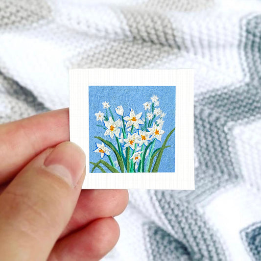 A hand is holding a small square card with a painting of white flowers against a blue background.