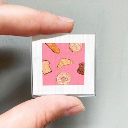A person's hand holding a small square with an illustration of various pastries, including a croissant, a donut, and a muffin, against a pink background.
