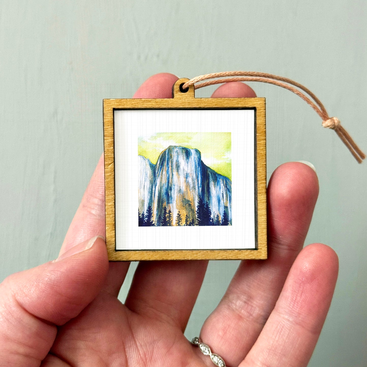 A hand holding a small wooden frame containing a watercolor painting of a mountain landscape.