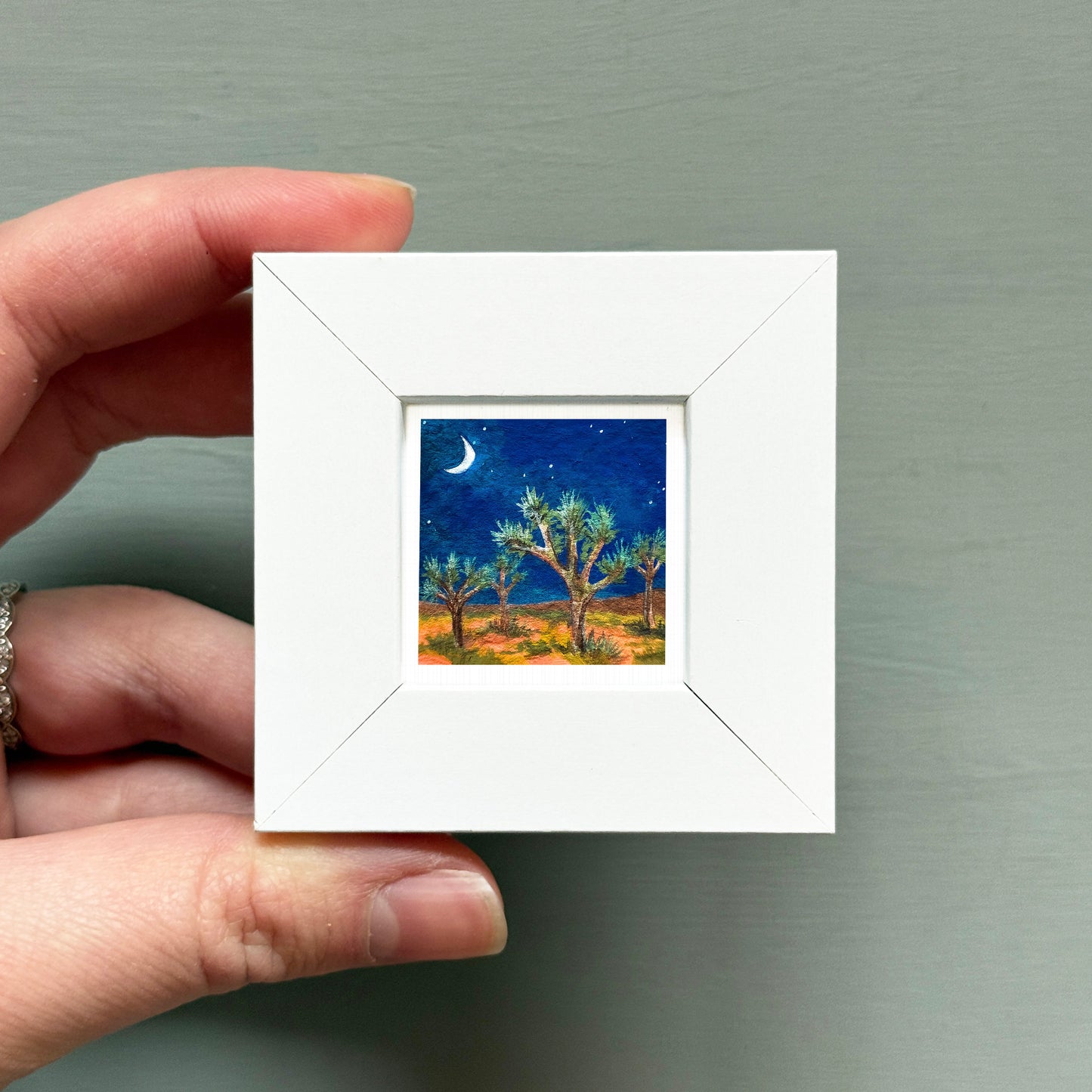 A hand is holding a small white square frame containing a painting of a desert landscape at night, with a crescent moon and stars in the sky.
