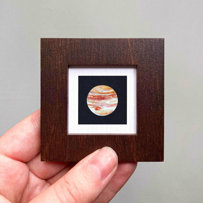 A hand is holding a small wooden frame containing a framed image of a planet.