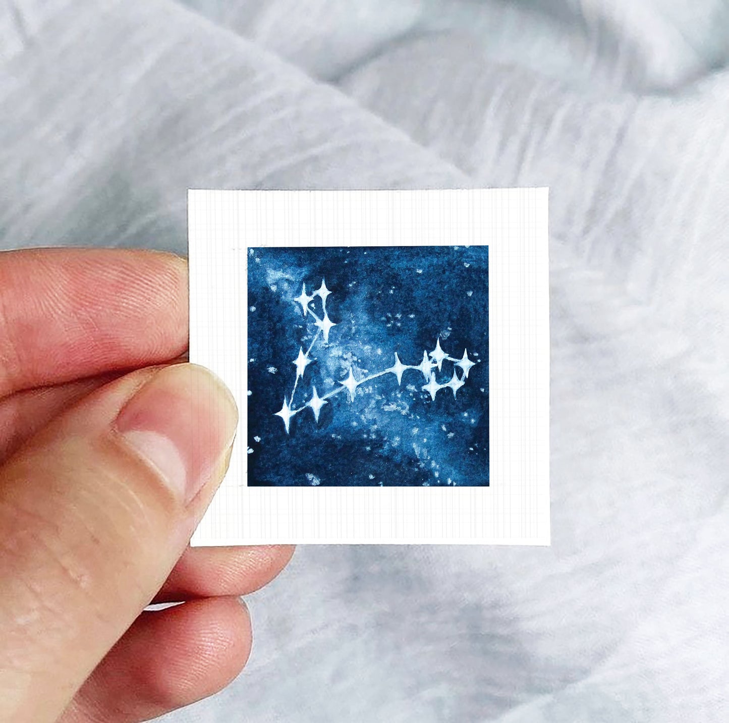 A hand is holding a small square-shaped paper with a depiction of a starry night sky on it.