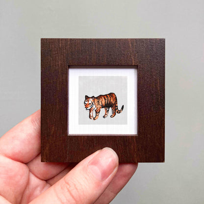 A hand is holding a small wooden frame containing a picture of a tiger.