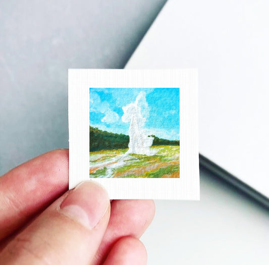 A hand is holding a small square painting depicting a landscape with a large geyser or fountain in the background.
