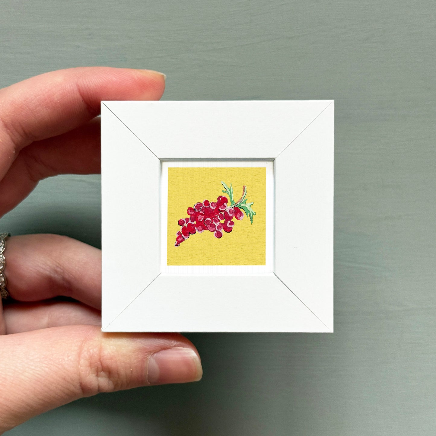 A hand is holding a small white frame containing a vibrant illustration of red grapes.