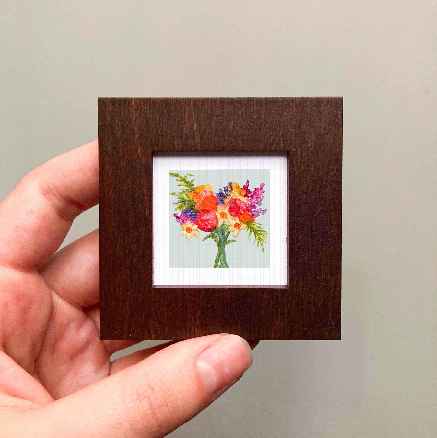 A hand is holding a small wooden frame containing a vibrant painting of a bouquet of colorful flowers.