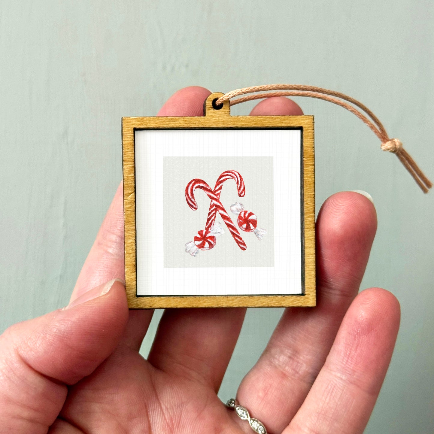 A hand holding a small wooden frame with a picture of two red and white striped candy canes inside it.