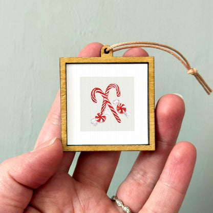 A hand holding a small wooden frame with a picture of two red and white striped candy canes inside it.