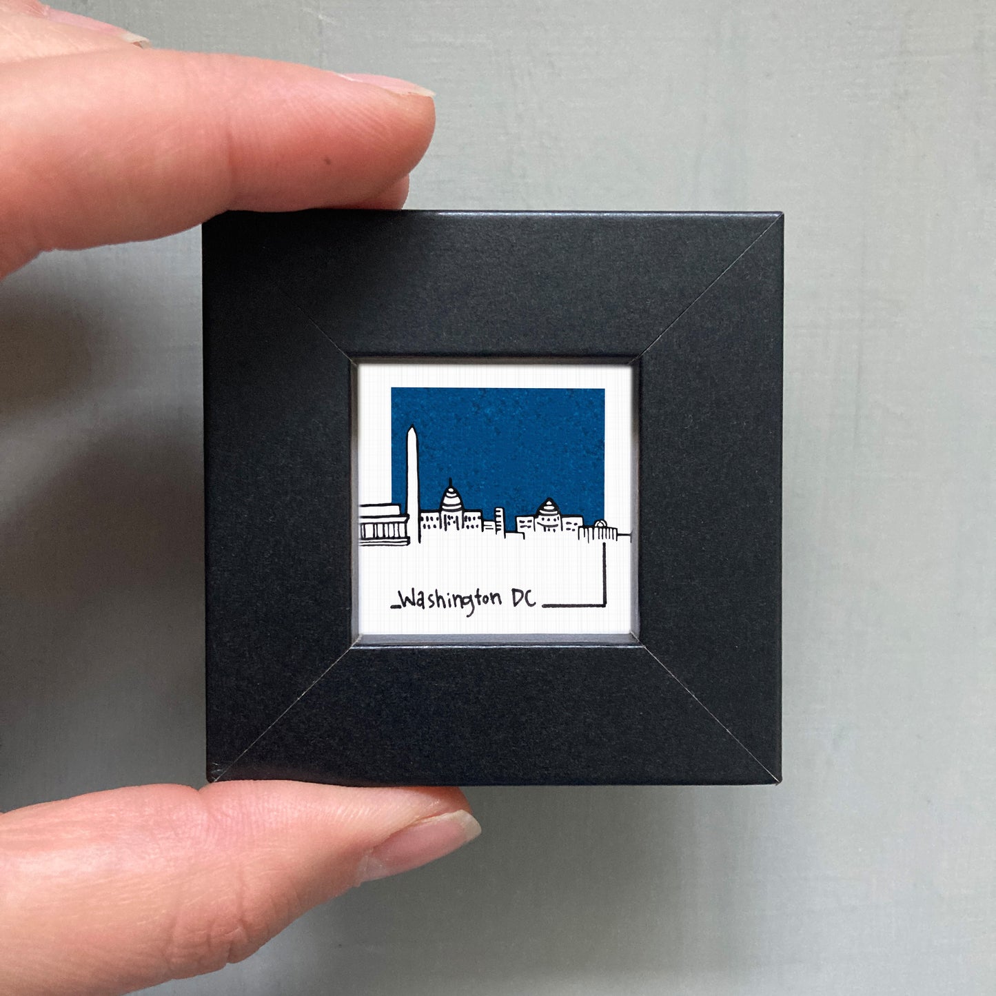 A hand is holding a small square frame containing an illustration of the Washington, D.C. skyline.