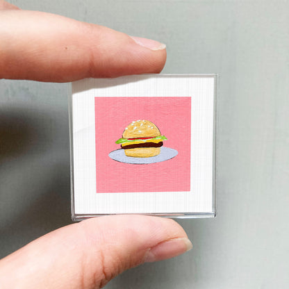 A person's hand is holding a small square with a picture of a hamburger on it.