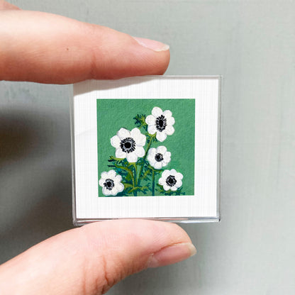 A person's hand holding a small square picture of white flowers against a green background.