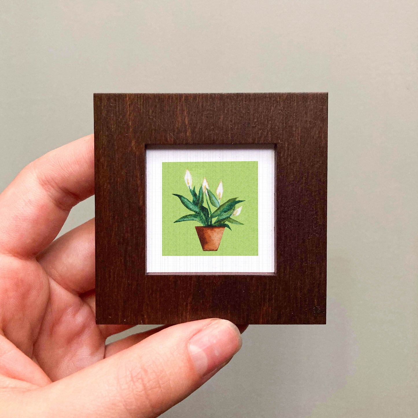A hand is holding a small wooden frame containing a framed picture of a potted plant with white flowers.