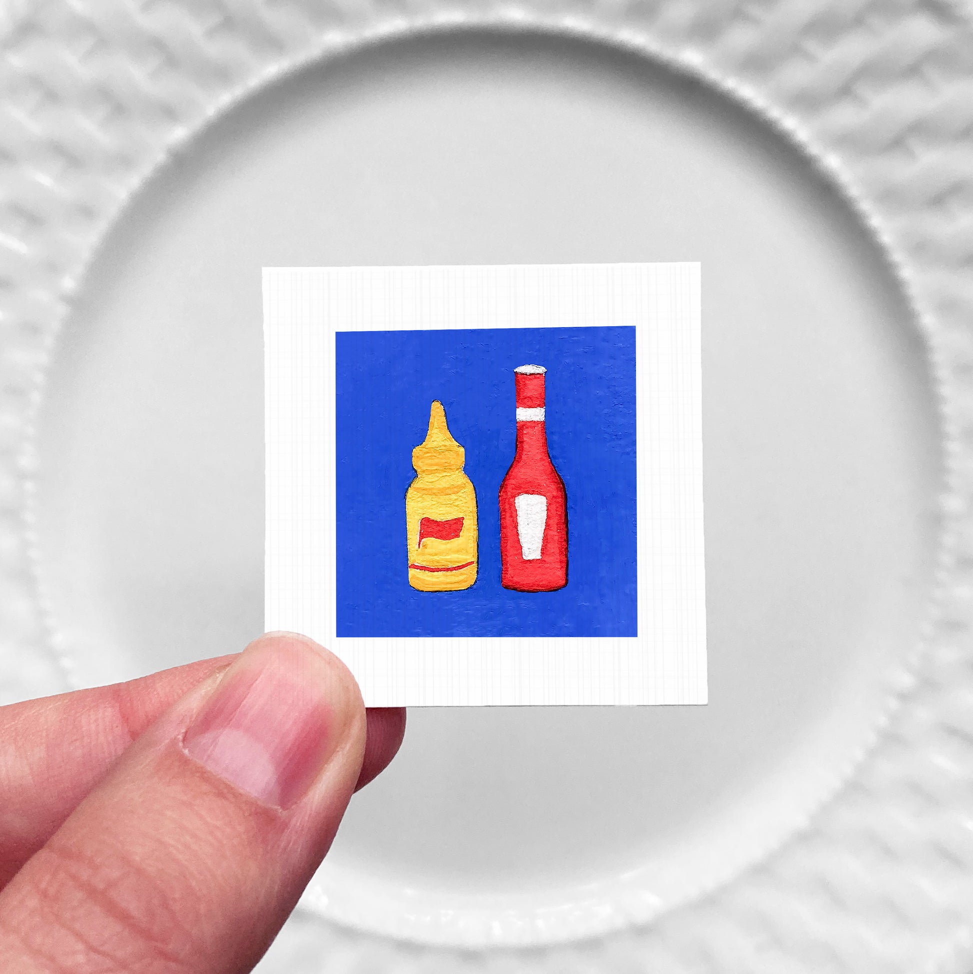 A hand is holding a small square with two bottles of condiments, one yellow and one red, against a white background.