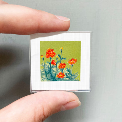 A person's hand is holding a small square picture of a vibrant flower arrangement against a green background.