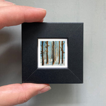 A person's hand holding a small black frame containing a painting of a snowy forest scene.