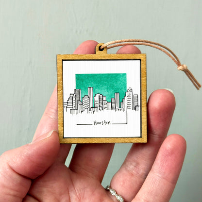 A hand holding a small wooden frame with a picture of a city skyline and the word "Houston" written on it.