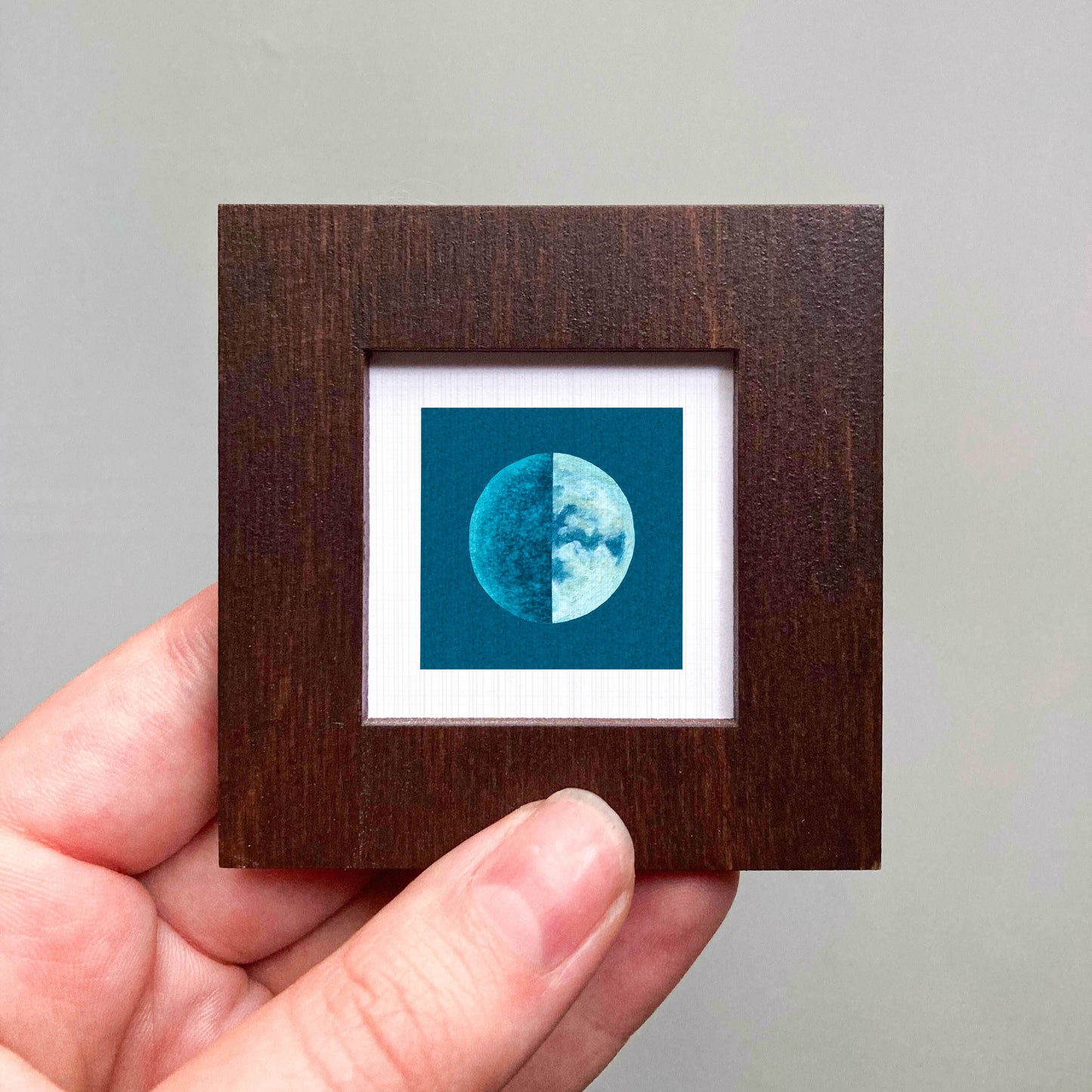 A hand is holding a small wooden frame containing a framed image of a blue and white crescent moon.