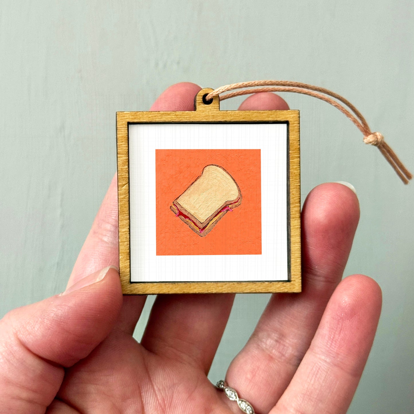 A person's hand holding a small wooden frame containing an illustration of a sandwich.