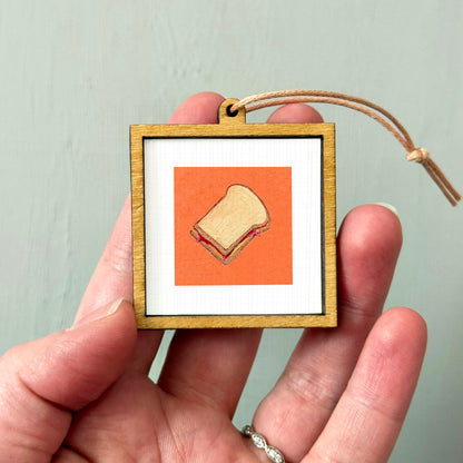 A person's hand holding a small wooden frame containing an illustration of a sandwich.