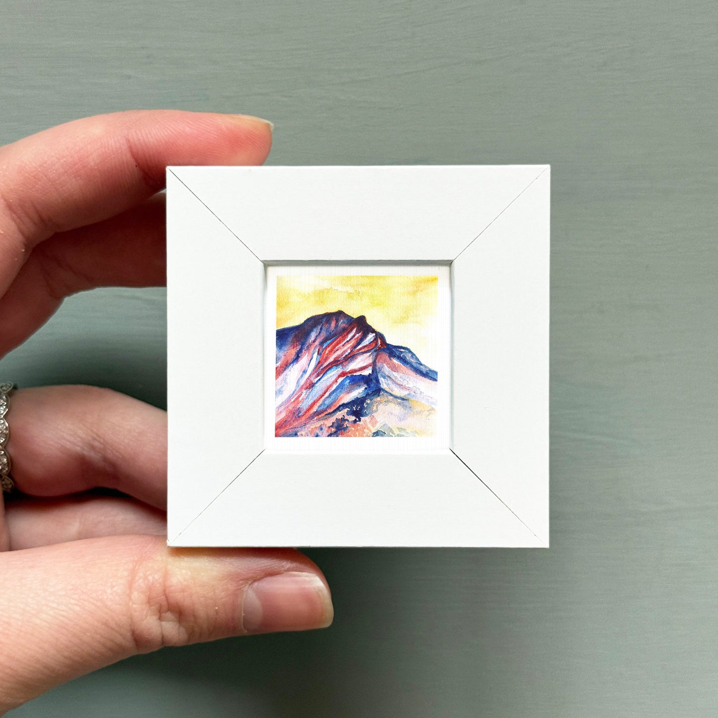 A hand is holding a small white square frame containing a colorful painting of a mountain landscape.