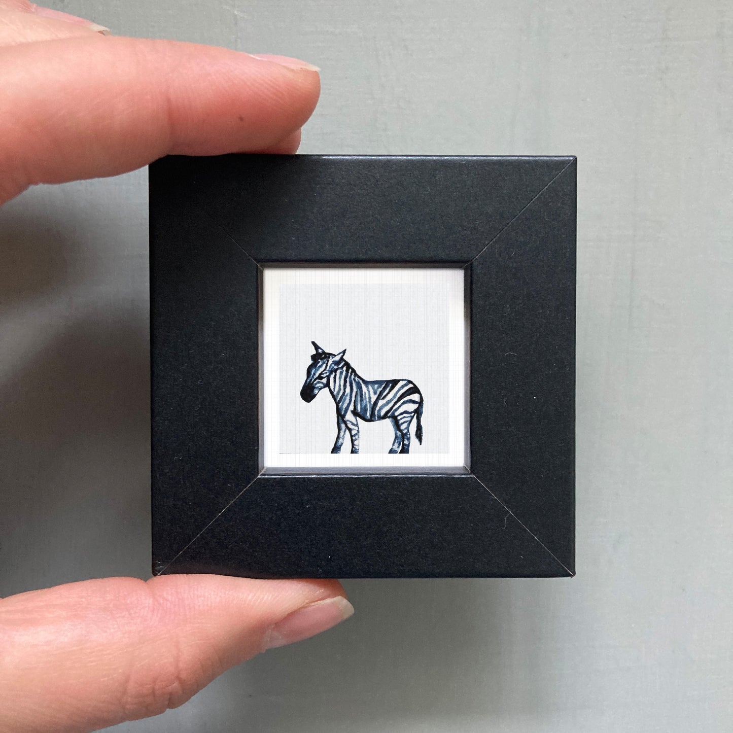 A person's hand is holding a small black frame containing a black and white illustration of a zebra.