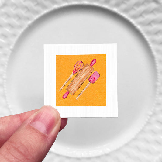 A hand is holding a small square with an illustration of a rolling pin and a pink spatula on it.