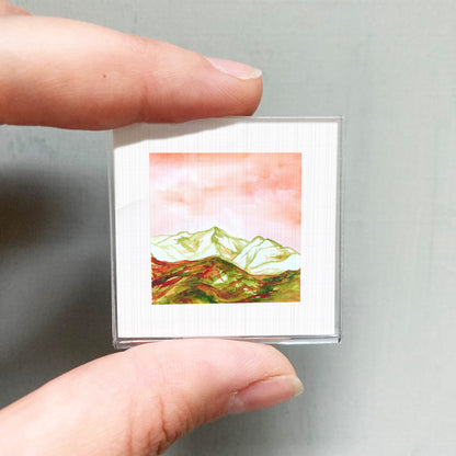 A person's hand is holding a small, square-shaped picture of a mountain landscape with a pink and green color scheme.