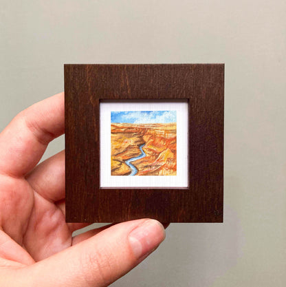 A hand is holding a small wooden frame containing a miniature painting of a desert landscape with a winding river.