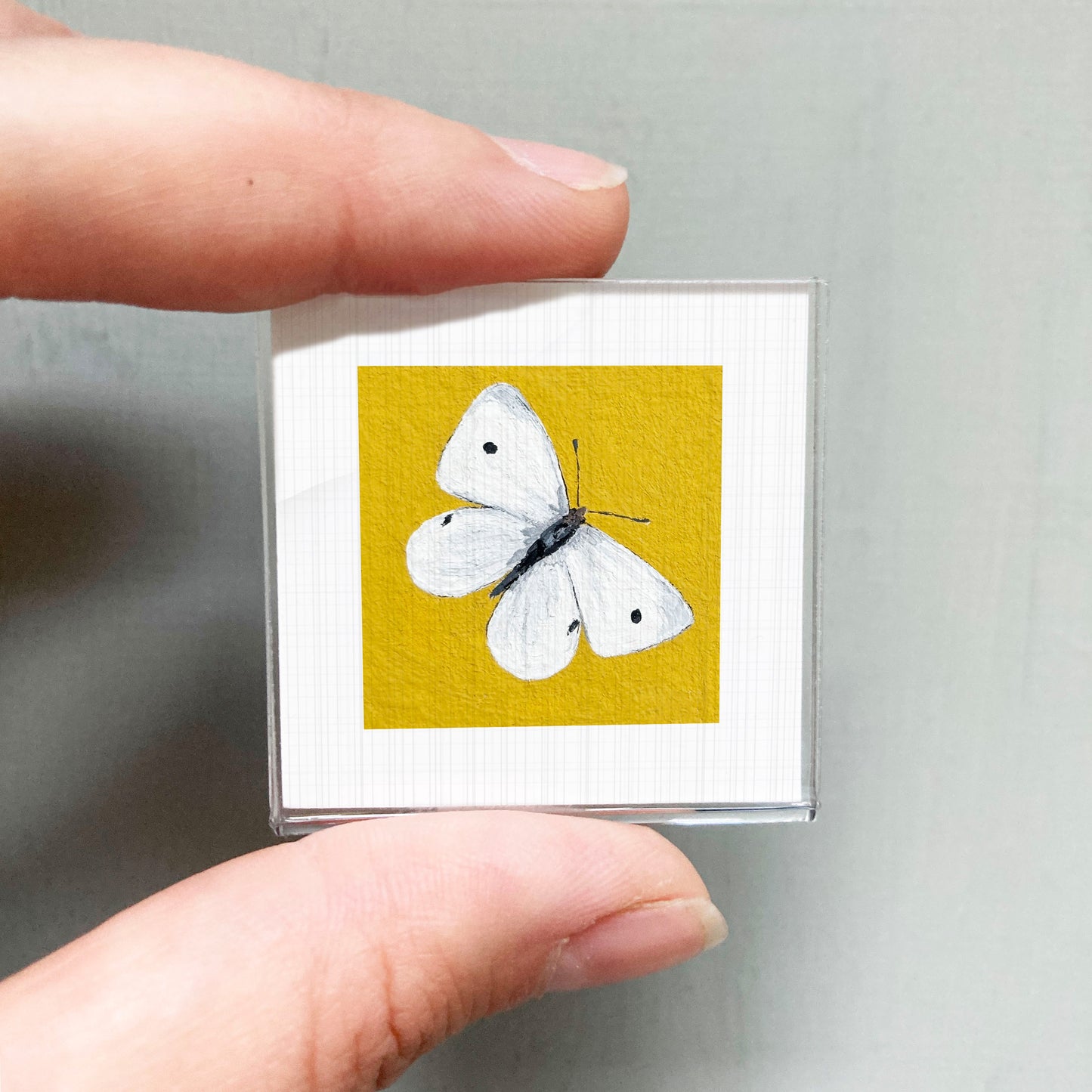 A hand is holding a small square with a white butterfly painted on it against a yellow background.